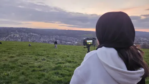 Aisha Khan/BBC A woman with a black headscarf and a white hoodie looks into a camera while standing on a green hill overlooking a city