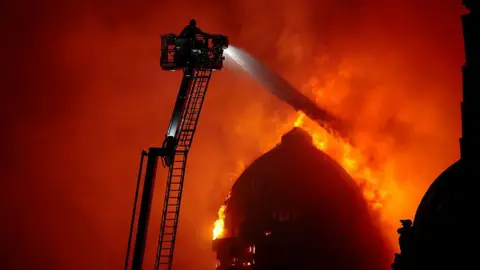 A firefighter on a large ladder sprays water over a burning domed building