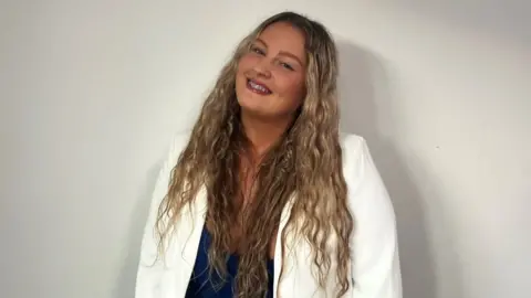 Mia Hardy Mia Hardy smiles at the camera. She has blonde, wavy hair, and is wearing a navy blue dress with a white blazer.