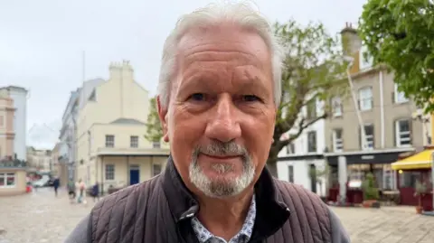Mick Tostevin is standing in Jersey's Royal Square. He is wearing a Oscar Jacobson body warmer.