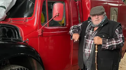 Richard Wallis is in a checked shirt and is wearing a black fleece waistcoat and a hat. He is stood next to a red vehicle.