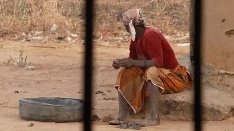 Sarah Peter sitting on a concrete slab. She is seen in the distance and photographed through the bars on a window. She is turning away from the camera.