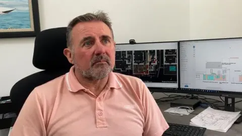 A middle aged man in a salmon coloured polo shirt looks to the right of the camera, sitting at a desk. behind him a computer screen shows architectual plans .