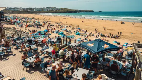 Boardmasters Festival / Adam Lievesley an aerial view of diners sitting at tables on a restaurant's decking, with parasols and umbrellas, in front of a golden beach full of beachgoers.
