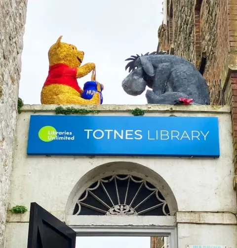 The photo is of two large sculptures above a doorway on a building. The children's book character Winnie the Pooh sits with a hand in his honey jar across from Eeyore the donkey. They are placed above a sign for Totnes Library, high above the entrance. 