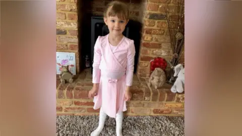 Family Photo Jessica in a pink ballet outfit, with brown hair, her hair tied back, standing in a living room in front of a brick fireplace.