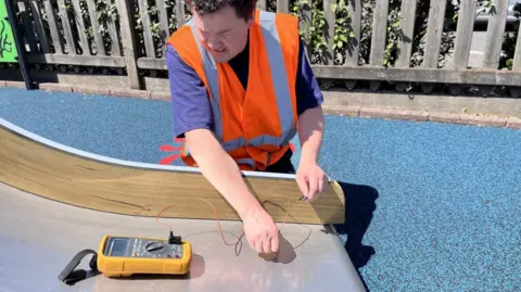 A man wearing a high vis vest sits on the playground floor at the bottom of a slide while holding a thermometer against its metal surface.