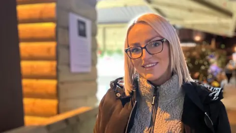 Jenna McCusker is standing in front of her market stall. 
She has blonde hair and is wearing black-rimmed glasses, half zip fleece and black coat.