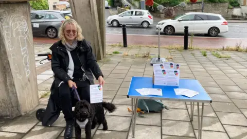 Brislington Welcome A woman sits on a chair with a black dog on a lead next to a table with leaflets. She is sitting beneath a flyover on a main road. 