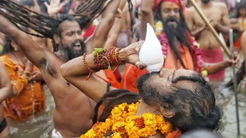 A sadhu wearing marigold garlands blows the conch as others around him take a dip in the river