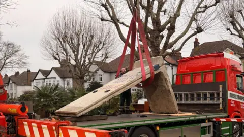 Steven Thorpe A red pick-up truck with a winch about the lift the piece of wall, parked by a row of houses