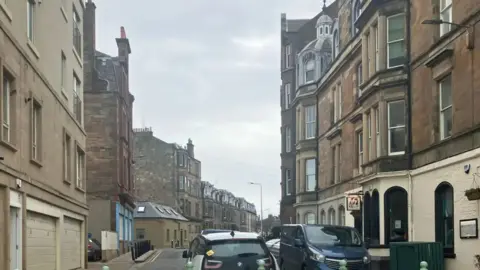 Bath Street in Portobello. Light-stone tenement buildings can be seen on either side. A white and black car is in the centre of the foreground. A dark-coloured van is parked on the right side of the road.