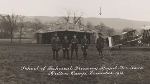 Handout A black and white picture of five airmen at an RAF base in December 1918, which is written in white at the bottom of the picture. A man is set aside from four other colleagues. An early plane is parked behind them.