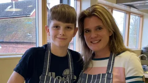 Ellen Roome and her son Jools Sweeney stand side by side in a large kitchen. They are both wearing dark blue aprons with thin white stripes on them. 
