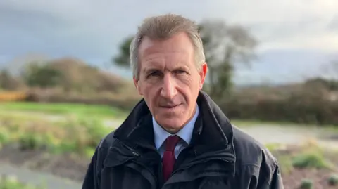 Dan Jarvis is looking serious and at the camera. There is a garden out of focus behind him. He has short grey hair and blue eyes. He is wearing a black waterproof jacket and a blue shirt with a burgundy tie.