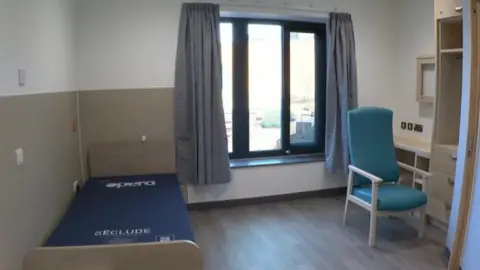 A hospital room with a blue medical bed, teal chair and light blue curtains. The floor is wooden and a large vertical window has light coming in.