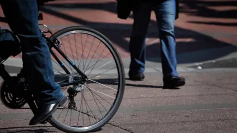 A generic image of a person riding a bicycle along a road while another person in the background walks across the road.