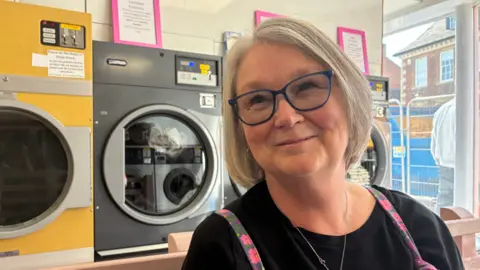 A woman in blue glasses wearing a black top smiles with washing machines behind her.