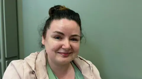 A women with dark hair staring at the camera. She is wearing a cream coat and a green t-shirt. Behind her is a green wall. 