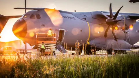 RAF The Atlas A400M on the runway, with staff bustling around it, and a side hatch open, with steps leading down. The sun is behind the plane, creating a golden lens flare.