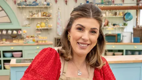 Love Productions Georgie Grasso smiling at the camera. She is wearing a red top underneath a beige apron. She is stood in the Bake Off tent. It is a head and shoulders shot of her. 