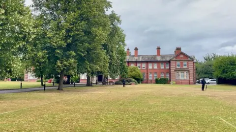 An ornate building with a large field in front of it as well as a playing field and trees. There is a row of trees to the left hand side of the image, obscuring part of the building. Two people in black clothing are standing in front of the building.