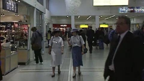 BBC Archive Two women walk through Terminal 1, they were white and blue uniforms, the pass under a large chandelier 