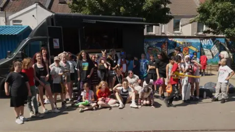 A group photo of children standing next to each other in front of a black van in the sun.