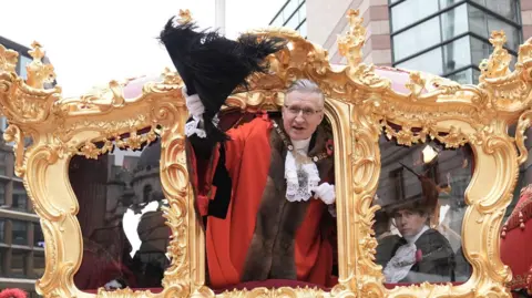 The Lord Mayor, dressed in ceremonial red robes with a fur collar and wearing a gold chain of office, leans out of the ornate gilded state carriage, raising a black feathered hat in greeting. The intricate golden frame of the carriage adds to the grandeur, and another official is visible inside, dressed in traditional attire.