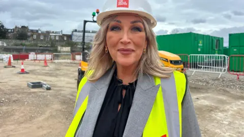 Deb Madden wearing a hard hat with The Skills Centre written on it. She is standing on a building site and is wearing a yellow hi-vis bib over a grey jacket and black shirt