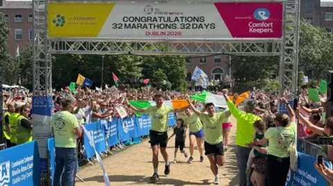 Jordan and Cian Adams crossing the finish line. They are both waving Irish flags. Above them is a banner saying "Congratulations 32 Marathons. 32 Days Dublin 2026".