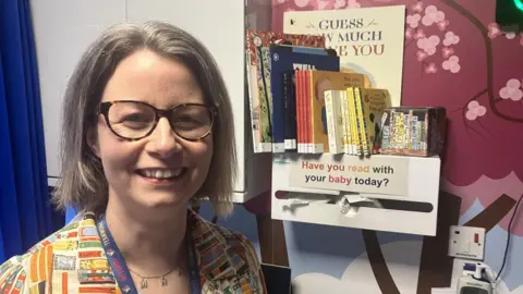 BBC/ Katie Radley A woman in a colourful top and wearing glasses is smiling at the camera. Behind her is a book shelf with children's books on and a sign reading "Have you read with your baby today?" and pink patterned wallpaper and a white cupboard