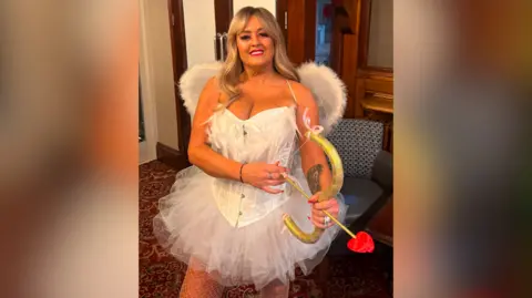 Lisa Hughes A blonde woman in a white tutu and angel wings with a red bow and arrow.