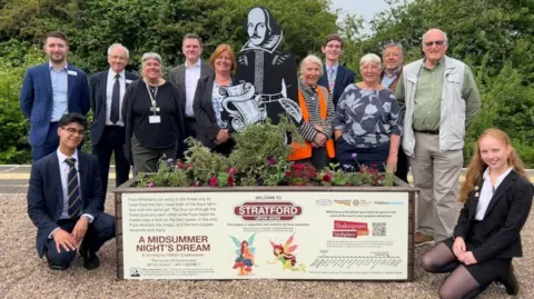 Network Rail A group of people standing on a train platform behind a raised bed with bedding plants in and a black and white statue of William Shakespeare in the middle.