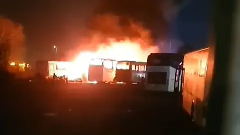 Large fire engulfing several buses in a dark outdoor lot at night, with thick black smoke rising into the sky.