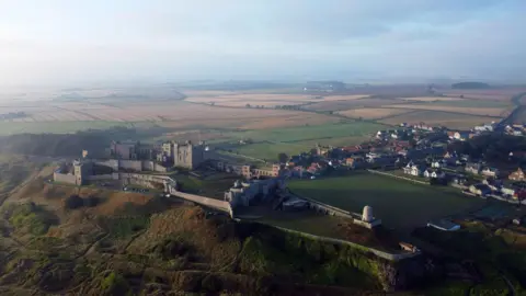 PA An aerial view of the village of Bamburgh with the castle in the foreground. It is a very large structure with a Norman keep in the centre and the walls clinging to the clifftop. The houses fill in behind the village green and in the distance fields stretch towards the horizon