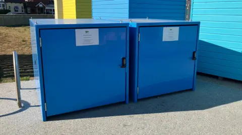 Jaimielee Rendall Two large blue metal containers next to a lighter blue beach hut.