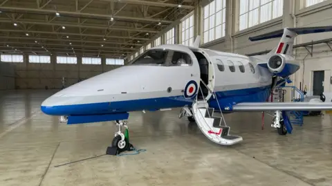 A small aircraft which is white, blue and red, is stationary inside a large warehouse building. It is being held to the spot by a small black wheel which has raised it from the ground, and steps on the side of it lie open. An RAF circular logo can be seen on its side.