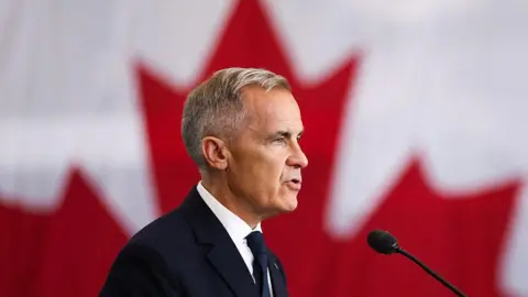 An image showing the side profile of Mark Carney, who is wearing a black suit and a black tie and is speaking into a microphone. He has short white and grey hair. Behind him is a blurred out Canadian flag, with the red maple leaf visible.