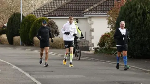 Three men running on a tarmac road while being followed by someone on a bicycle