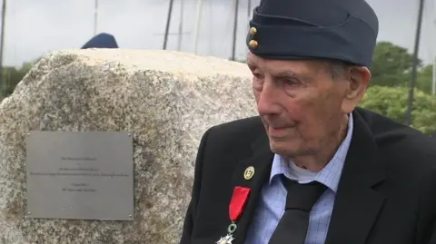 D-Day veteran Albert Westgate wearing a blue suit and medal with red ribbon, sitting beside the granite memorial