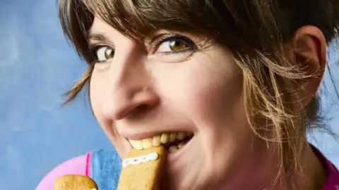 Tat Effby A woman with short brown hair and a biscuit between her teeth in front of a blue background