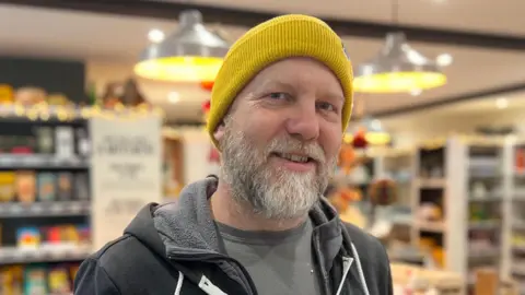BBC Jock Pettitt pictured wearing a yellow beanie and black zip-up hoodie. He has a grey and white beard and is smiling at the camera. 