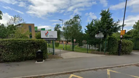 Google A school building surrounded by trees and hedges. It is a sunny day.