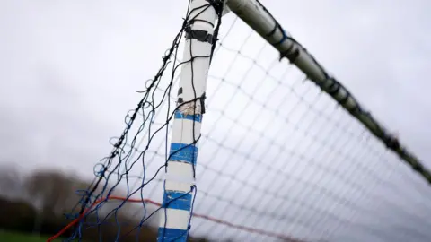A close up of a tatty football goal post and the net behind it. The photo is shot upwards towards the grey sky.