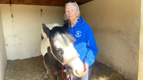Karen Thompson, who has white hair and is wearing a blue fleece top, stands next to a black and white pony