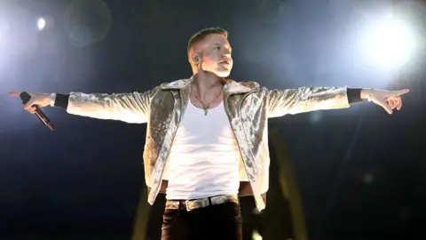Getty Images Macklemore, dressed in a white under-vest and metallic gold jacket, stretches his arms out to either side in recognition of the crowd before the NRL grand final in Sydney