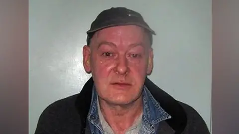 A custody-style photograph showing a Sweeney wearing a dark cap, denim shirt layered under a dark jacket and a light-coloured top, standing against a plain light background.