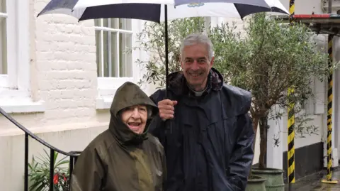 STFH Phyll with reverend Canon Edward Probert from Salisbury Cathedral standing in rain coats on a street underneath an umbrella