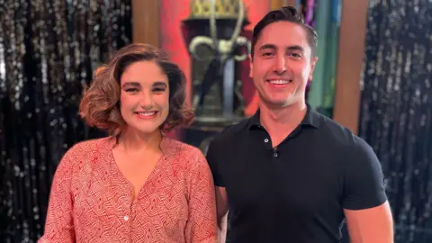 BBC Natasha is in the red patterned dress and has curled her hair. Connor Carson is standing beside her in a tight black t-shirt. There are standing on the stage and some of the set is visible behind them